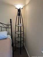 Full view of black metal torchiere floor lamp with frosted glass shade turned on, positioned next to bed.