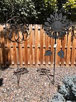 Two metal flower wind spinners standing in a backyard garden bed with rock ground cover and wooden fence background