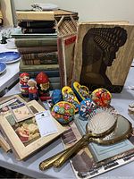 Photo showing a large assortment of vintage books stacked, wooden nesting dolls, multiple colorful metal hand-held noise makers, vintage gold-toned hairbrush with mirror, and some vintage paper booklets