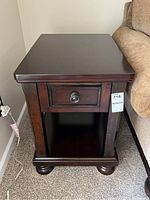 Front view of the dark stained wood side table next to a couch showing drawer, bun feet and bottom shelf.