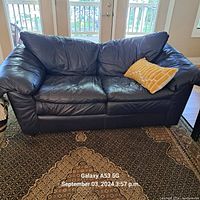 Photo of a black leather loveseat with cushioned seats and armrests, showing its condition and included decorative pillow.
