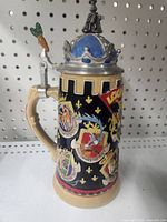 Front and left side view of Looney Tunes collectible stein showing colorful character crests and beige handle with carrot lever on metal lid.