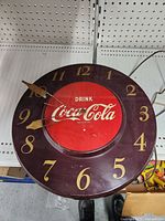 Top view of a round Drink Coca Cola wall clock with red center and gold numbers on dark background.