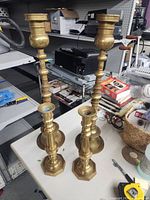 Four brass candle sticks, 2 pairs, 25in and 14in tall arranged on table with other items in background.