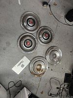 Wide view of five metal hub caps and one white Scarab vanity plate on concrete floor showing wear and dirt.