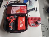 Milwaukee cordless drill and battery pack inside open carrying case with charger unit next to it on a table