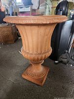 Side view of one terracotta-style flower urn showing fluted body, scalloped wide rim, pedestal base and square plinth base.