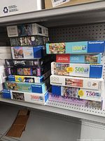 Shelf with a large assortment of puzzle boxes stacked including multiple Ravensburger jigsaw puzzles of various piece counts (500, 1000, 2000, 3000) and boxes from other brands like Clementoni.