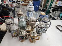 Full lot showing 11 beer steins with a mix of ceramic and glass bodies and pewter lids.