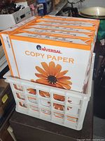 Seven unopened packages of Universal Copy Paper stacked in white crate, displaying packaging with daisy flower.