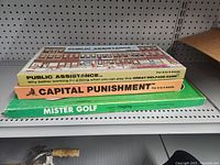 Three vintage board games stacked on a shelf with visible titles and artwork: Public Assistance, Capital Punishment, Mister Golf.