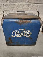 Front view of vintage blue Pepsi-Cola cooler showing logo and rust spots on metal surface.