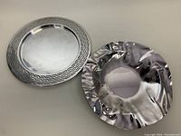 Pair of stainless steel serving trays, SHAMAN platter with geometric cutout rim and polished freeform VENTO ALEX tray, side by side from top view.