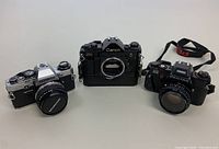 Three 35mm SLR cameras side by side: silver Olympus OM-10 with lens, black Canon A-1 body, black Pentax Program Plus with lens