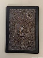 Full view of the antique silver plaque/book cover showing embossed religious figures and ornamental border on dark frame.