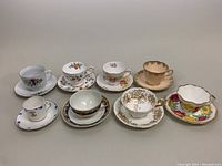 All seven demitasse teacup and saucer sets arranged on table showing different patterns and colors including floral and gold trim