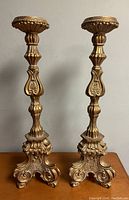 Full view of the pair of tall carved pillar candleholders showing overall shape, gold finish, and ornamental base and shaft details.