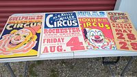 Four vintage circus advertisement posters displayed on a table outdoors, showing general condition and sizes.