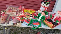 Wide view of vintage Christmas decorations and paper advertising items on a table outdoors, showing Santa sleigh, elf doll, snowman figure, and paper signs.