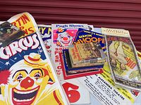 Photo showing several vibrant circus posters including a clown with a large red nose and yellow hat, and a small stack of colorful circus-related books on top of various posters.