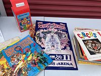 Photo of metal Rocket Ride Carousel toy in red box, bamboo clown figurine, and several circus books and posters arranged on a table.