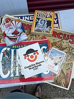 Photo showing collection of circus memorabilia including colorful posters featuring lions and circus acts, and several books with circus and clown themes.