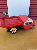 Side view of red, black, yellow, and white metal friction dump truck toy on yellow surface with white original box in background.