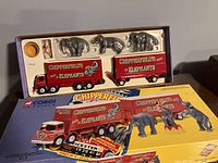 Full box set showing red lorry and trailer, three elephant figurines, rider figure, and pedestal with packaging and promotional materials.
