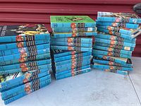 Multiple stacks of Hardy Boy books mostly blue hardcovers with black labels and illustrations on spine, arranged in multiple stacks on floor.