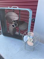 Large blue Electrohome branded floor fan with rusted metal grill and blades, and small green and pink vintage table fan on a blue surface.
