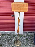 Full view of the vintage floor lamp standing outdoors, showing the woven wicker lampshade, brass pole, wooden decorations, and round base.