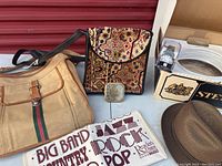 Photo showing beige canvas tote bag with handles, ladies tapestry handbag with floral pattern, ladies compact, wristwatch in packaging, and a sign with music genres