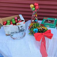Wide view showing entire lot on white cloth including holly tree, glass vase with ornaments, box of Christmas balls, Christmas tree top in box, and glass dish with handle.
