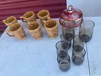 Full lot view showing five ceramic mugs, seven taupe glasses (two highball and five juice), and the cranberry red covered candy dish, displayed on a white surface.