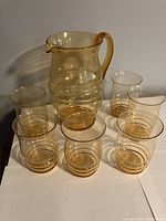 Full set of one pitcher and six glasses shown together on white surface in ambient light.