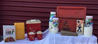 Wide shot of all items showing metal bread box, flour bucket, salt and pepper shakers, napkin dispenser, Kellogg's Corn Flakes tin, Cracker Jack box, multiple collectible plastic cups, and cork stoppers.