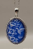 Close-up of oval blue resin cameo pendant set in sterling silver with a detailed white hummingbird and floral design, bordered by small Swarovski crystals.