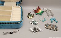 Blue velvet zippered storage box and assortment of vintage sterling silver brooches and earrings displayed on neutral surface, showing variety including enamel leaf and abalone fish designs with turquoise earrings.
