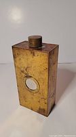 View of rectangular weathered coppery-bronze flask with circular reflective inset, showing front and side with visible tarnish and patina.