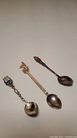 Photo showing three silver souvenir spoons lying flat with detailed decorative handles visible.