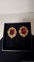 Pair of gold-tone clip-on earrings with a large red rectangular crystal center surrounded by smaller amber and cranberry crystals, displayed in a black box.