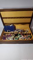 Top view of open wooden jewellery box showing large collection of mixed jewellery items and a blue sparkly purse