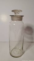 Full view of clear glass apothecary bottle standing upright showing cylindrical shape, clear body, and glass stopper.