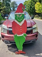 Front view of the tall wooden cutout painting of the Grinch character in Santa outfit with bell on hat, placed in front of a red vehicle.