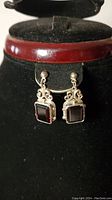 Front view of sterling silver earrings with rectangular ruby red stones displayed on black velvet stand with maroon trim