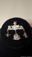 Front view of silver brooch showing cat with book and three hanging charms shaped as teapots and a cup inscribed 'Cup Of Kindness'.