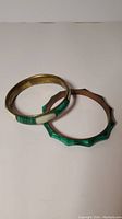 Photo showing two bracelets: a smooth brass bangle with malachite and white inlays, and a copper bangle with scalloped edges and malachite inlay.
