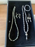 Two handcrafted jewelry sets displayed on a black tray. The left set has a serpentine jasper bead necklace and matching earrings. The right set has a mauve pearl necklace, bracelet, and matching earrings with sterling silver ear wires.