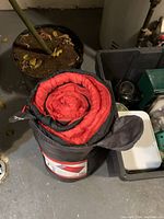 Red and black sleeping bag rolled in a storage bag placed on the floor near a plant.