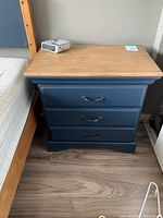 Full view of night stand showing painted blue body, natural wood tabletop, three drawers with new handles, placed next to bed.